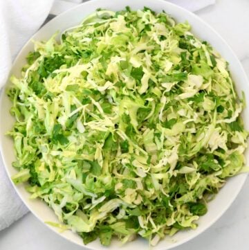 A large white bowl filled with finely shredded green cabbage and cilantro, placed on a white surface with part of a white cloth visible in the background.