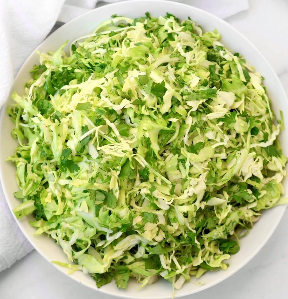 A large white bowl filled with finely shredded green cabbage and cilantro, placed on a white surface with part of a white cloth visible in the background.