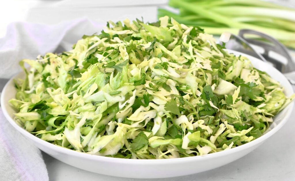A white bowl filled with freshly shredded cabbage, cilantro, and green onions, sitting on a white surface with a cloth and whole green onions in the background.