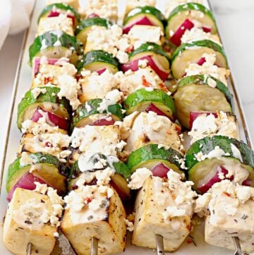 Skewers of grilled tofu, zucchini, and red onion topped with crumbled feta cheese and herbs create a vibrant tofu souvlaki, neatly arranged on a white rectangular platter.