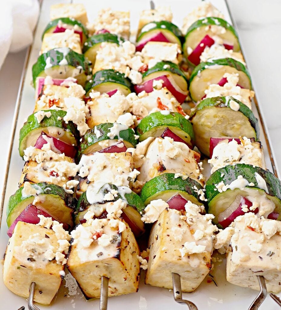 Skewers of grilled tofu, zucchini, and red onion topped with crumbled feta cheese and herbs create a vibrant tofu souvlaki, neatly arranged on a white rectangular platter.