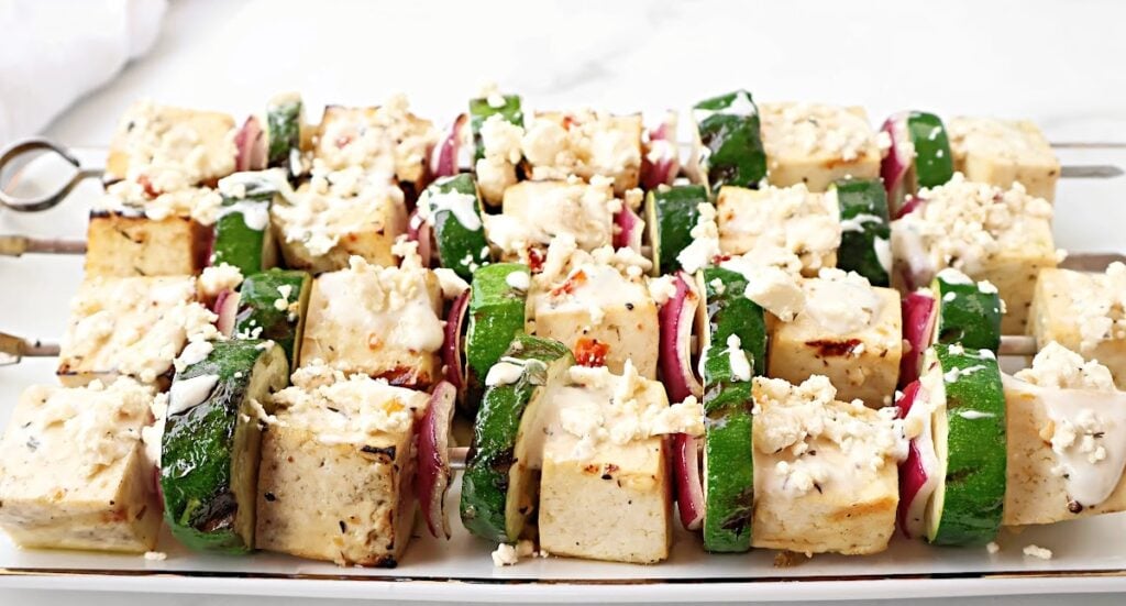 Skewers with grilled tofu cubes, zucchini slices, and red onion pieces are arranged on a white platter in this vibrant Greek tofu recipe, topped with a sprinkle of crumbled cheese or dressing.